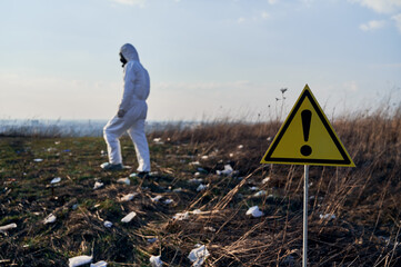 Focus on warning sign. Back view of male ecologist in protective suit walking in field with trash...