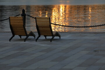 Wooden chairs on the embankment by the water with a view of the reflection and glare of the rising...