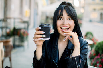 Happy excited emotional young Asian brunette woman sitting at outdoor urban cafe with a cup of coffee and looking at camera with smile. Chinese girl spending free time at outdoor cafe.