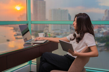 Asian businesswoman drinking coffee working on new project on digital tablet with laptop in background of river beautiful view at sunset and background blur building skyscrapers.