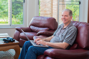 Portrait of elderly man sitting on sofa, holding laptop computer, smiling happily at camera and home interior on background