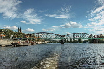 Naklejka premium Postcard view of Vysehrad standing on massive rock looming over the Vltava river,Prague,Czech Republic.Czech national cultural monument.Riverfront houses,bridge,boats on sunny day.Sailing on river.