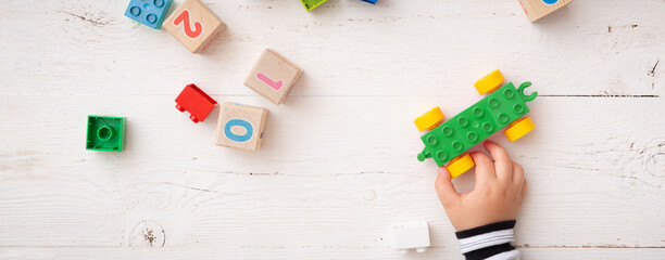 A wide view of children's hands playing with children's educational toys, green car, colorful cubes, bricks and wooden cubes with numbers on a white wooden background. Concept development
