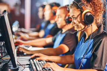 Side view of a happy mixed race girl, female cybersport gamer wearing headphone sitting in one row with team, participating in esports tournament