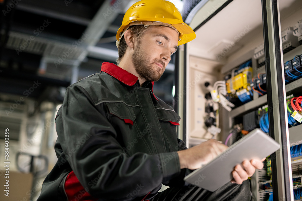 Poster young serious engineer in workwear and hardhat scrolling through technical data