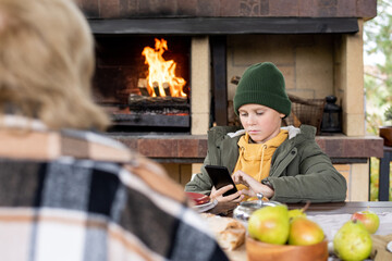Serious clever schoolboy in beanie and warm jacket scrolling in smartphone