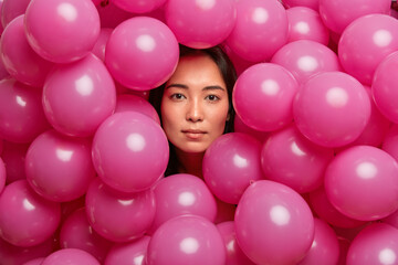 Fototapeta na wymiar Serious confident brunette Asian woman looks through air balloons has confident gaze at camera prepares for holiday celebration makes photo near decorated background. Birthday party concept.