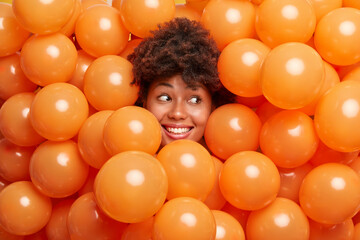 Cheerful curly haired ethnic woman celebrates birthday and surrounded by orange helium balloons looks away with broad smile has fun on party poses over decorated background. Holiday concept.
