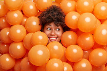 Cheerful curly haired ethnic woman celebrates birthday and surrounded by orange helium balloons...