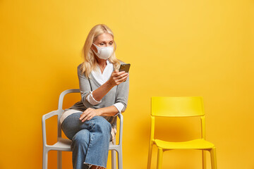 Serious mature woman infected by coronavirus wears medical mask and concentrated in smartphone feels bored lonely during self isolation at home sits on comfortable chair surfs social networks