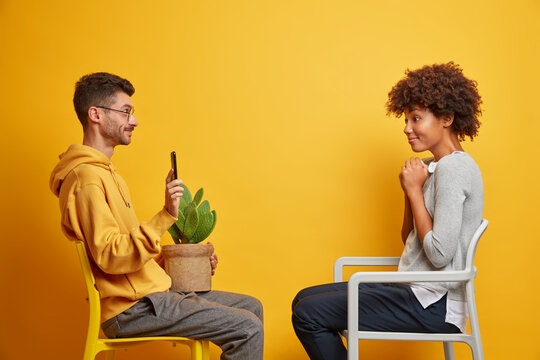 Horizontal shot of handsome boyfriend sits on comfortable chair opposite girlfriend shows some photos on mobile phone carries pot with cactus spend free time at home isolated over yellow background