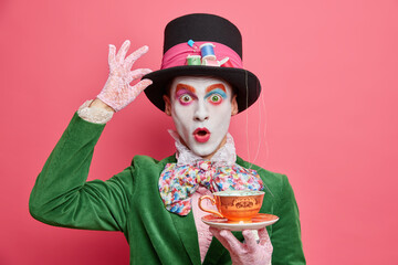 Surprised male hatter wears gentlemans clothes holds cup of hot tea pretends to come from...