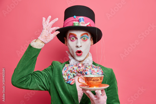Surprised male hatter wears gentlemans clothes holds cup of hot tea pretends to come from wonderland wears vivid makeup celebrates halloween on costume party poses indoor alone. Body painting