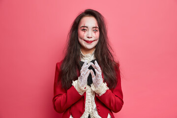 Happy vampire woman has devil plan and intention to do something wears halloween makeup and masquerade costume poses indoor against rosy background. Terrifying female has image of spooky zombie - obrazy, fototapety, plakaty