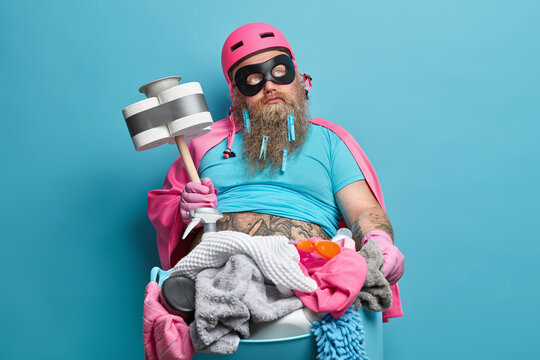 Handsome bearded adult man with thoughtful expression does housework and poses with basin full of laundry holds cleaning tool dressed in superhero costume poses indoor against blue background