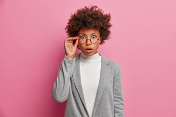 Fototapeta na wymiar Indoor shot of embarrassed dark skinned woman stares through glasses being amazed by awful news dressed in formal grey outfit isolated on pink background. Shocked terrified female entrepreneur