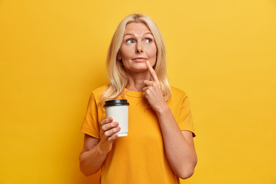 Photo of thoughtful wise fifty years old woman concentrated somewhere with pensive expression drinks coffee to go dressed in casual t shirt makes plans in mind isolated on yellow background.