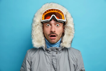 Close up shot of embarrassed active man enjoys favorite winter sport and stares with shocked expression at camera dressed in warm outerwear poses indoor. Recreation season lifestyle concept.
