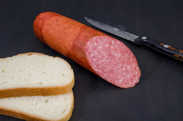 half a stick of sausage, two slices of bread and a knife on a black wooden table. Delicious salami
