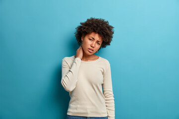 Fototapeta na wymiar Displeased Afro American woman touches neck suffers osteochandrosis leads sedentrary lifestyle feels strong pain dressed in casual white jumper isolated on blue background looks with unhappy grimace