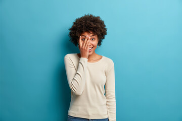 Fototapeta na wymiar Photo of cheerful optimistic joyful Afro American woman hides face with hand looks through fingers and wears casual jumper foolishes around poses against blue background. Happy emotions concept