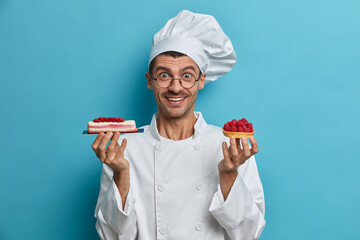 Positive professional confectioner holds tasty handmade desserts with berries wears white uniform...