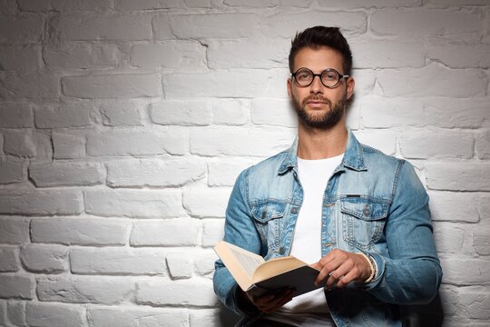 Smart casual man in glasses reading book, leading against brick wall. Copyspace.