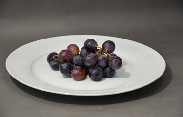 Round black grapes on a white plate. Useful fruits on a gray background. Organic grapes on a branch.
