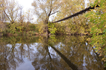 Photo of a beautiful river with a bridge made of wood. Reflection in the water of autumn trees and a bridge. Autumn landscape of the river. Water with leaves. Yellow leaves. Circles on the water