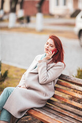 A beautiful red-haired girl sits on a park bench with a phone on her hand. Modern communications. Distance work and education.