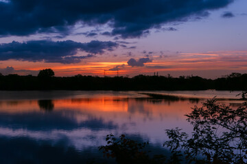 Amazing sunrise in rural scene. Symmetry of the sky in a lake at sunset. Clouds reflecting on the water. Quiet relaxing scene with a beautiful colorful cumulonimbus. Silhouette of vegetations.