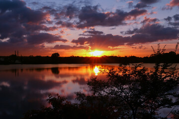 Amazing sunrise in rural scene. Symmetry of the sky in a lake at sunset. Clouds reflecting on the water. Quiet relaxing scene with a beautiful colorful cumulonimbus. Silhouette of vegetations.