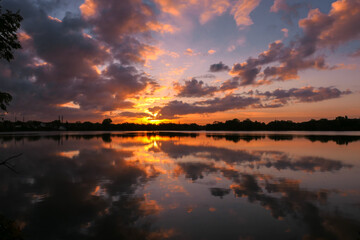 Fototapeta na wymiar Amazing sunrise in rural scene. Symmetry of the sky in a lake at sunset. Clouds reflecting on the water. Quiet relaxing scene with a beautiful colorful cumulonimbus. Silhouette of vegetations.
