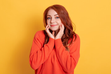 Shy delightful pleased female keeps hands on cheeks, looks with love and sympathy at camera, beautiful ginger girl wearing orange warm orange jumper against yellow wall.