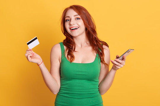 Image of beautiful red haired woman wearing green dress holding credit card and cellphone, looks at camera with excited expression, red haired female with unlimited bank card.