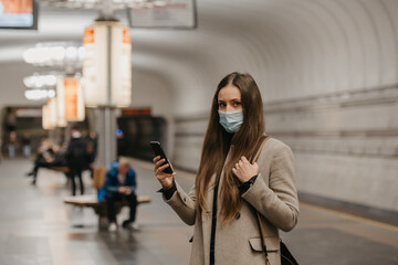 A woman in a face mask to avoid the spread of coronavirus is posing with a smartphone at a subway station. A girl in a surgical face mask against COVID-19 is waiting for a train on a metro platform.