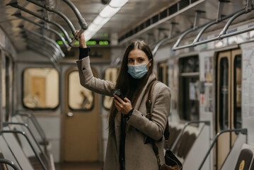 A woman in a medical face mask to avoid the spread of coronavirus is posing in a subway car. A girl with long hair in a surgical mask on her face against COVID-19 is holding a cellphone on a train.