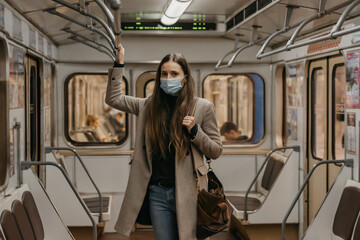 A woman in a face mask to avoid the spread of coronavirus is holding onto the handrail and posing in the center of the subway car. Girl in a surgical mask against COVID-19 is standing on a metro train