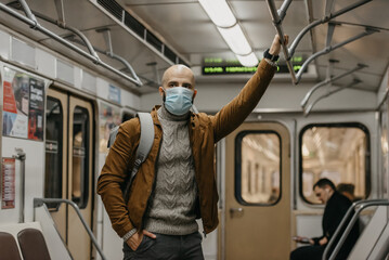 Fototapeta na wymiar A man with a beard in a face mask to avoid the spread of coronavirus is holding onto the handrail in a subway car. A bald guy in a surgical face mask against COVID-19 is standing on a metro train.
