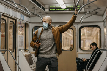 A man with a beard in a medical face mask to avoid the spread of coronavirus is looking around in a subway car. A bald guy in a surgical mask against COVID-19 is holding a cellphone on a metro train.