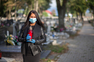 A woman wearing a protective mask against the coronavirus COVID-19 SARS-CoV-2 holds a candle in her hand and visits her relatives at the cemetery. Lockdown cemetery