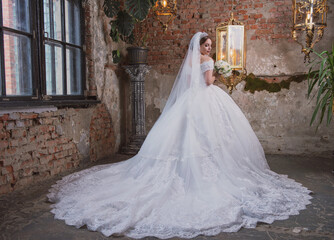 bride in a wedding dress with a long train and a bouquet in an old hall with chandeliers - 384429171