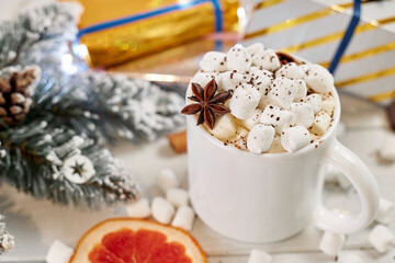Mug of hot coffee with marshmallows sprinkled with chocolate, anise, cinnamon and a slice of dried grapefruit on white wooden table. Christmas concept. Winter hot drink and gift boxes.