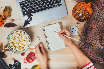 Top view woman writing plans or list of films for Halloween movie party. Blank notebook, laptop, popcorn bowl, pumpkin, bat, ghost, fall leaves, warm plaid on wooden background. Cozy and safe holiday.