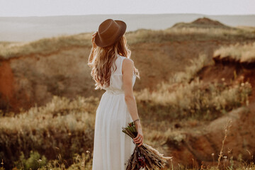 the bride walks on the field in the rays of the setting sun, the bride's white dress glows in the rays of the sun, the bride in a boho style hat, the concept of a wedding abroad