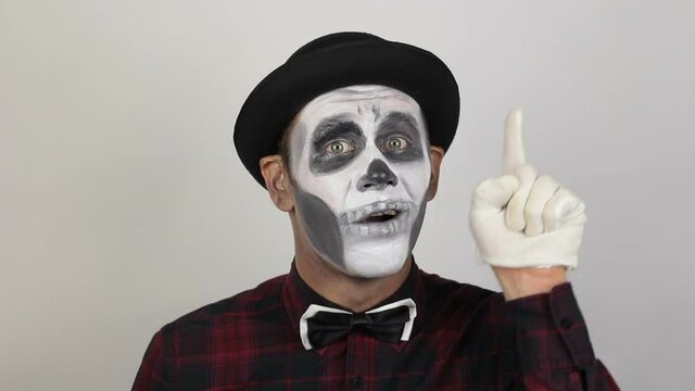 Terrible man in clown makeup, pointing up with his index finger. The scary clown looks at the camera and laughs terribly and points up with his index finger. Halloween costume.