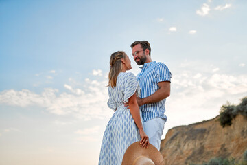 A loving young couple hug on the seashore, enjoying each other and their vacation. A woman in a dress and a man in a shirt and white trousers are walking on the beach.
