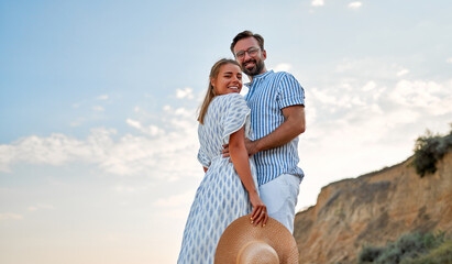 A loving young couple hug on the seashore, enjoying each other and their vacation. A woman in a dress and a man in a shirt and white trousers are hugging on the beach.