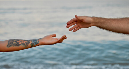 Close-up cropped image of the hands of a young couple against the backdrop of the sea and waves that stretch towards each other.