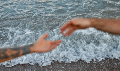 Close-up cropped image of the hands of a young couple against the backdrop of the sea and waves that stretch towards each other.